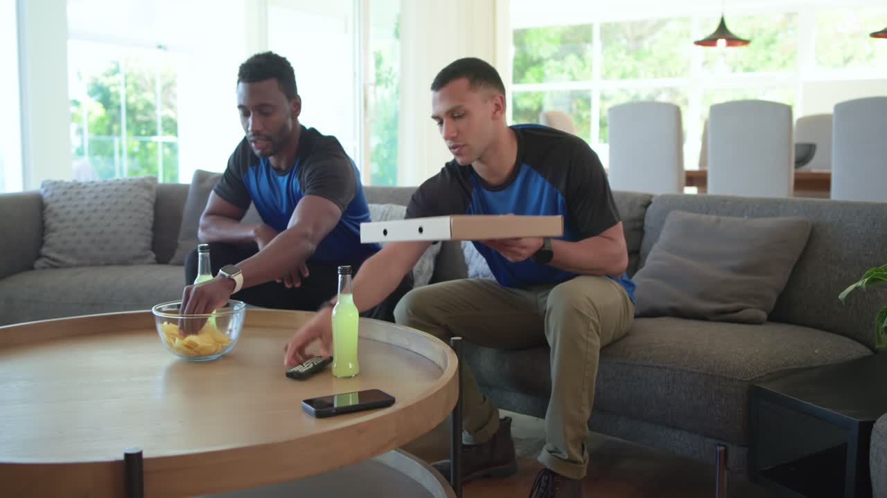 Diverse male friends entering lounge carrying pizza, chips, opening box grabbing snacks watching TV