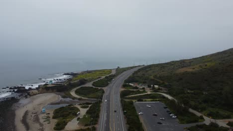 car driving on a road trip up the california coastline on a cloudy day. the lush green hills contrast the sandy beach and the black rocks jutting out of the pacific ocean. 4k drone travel shot.