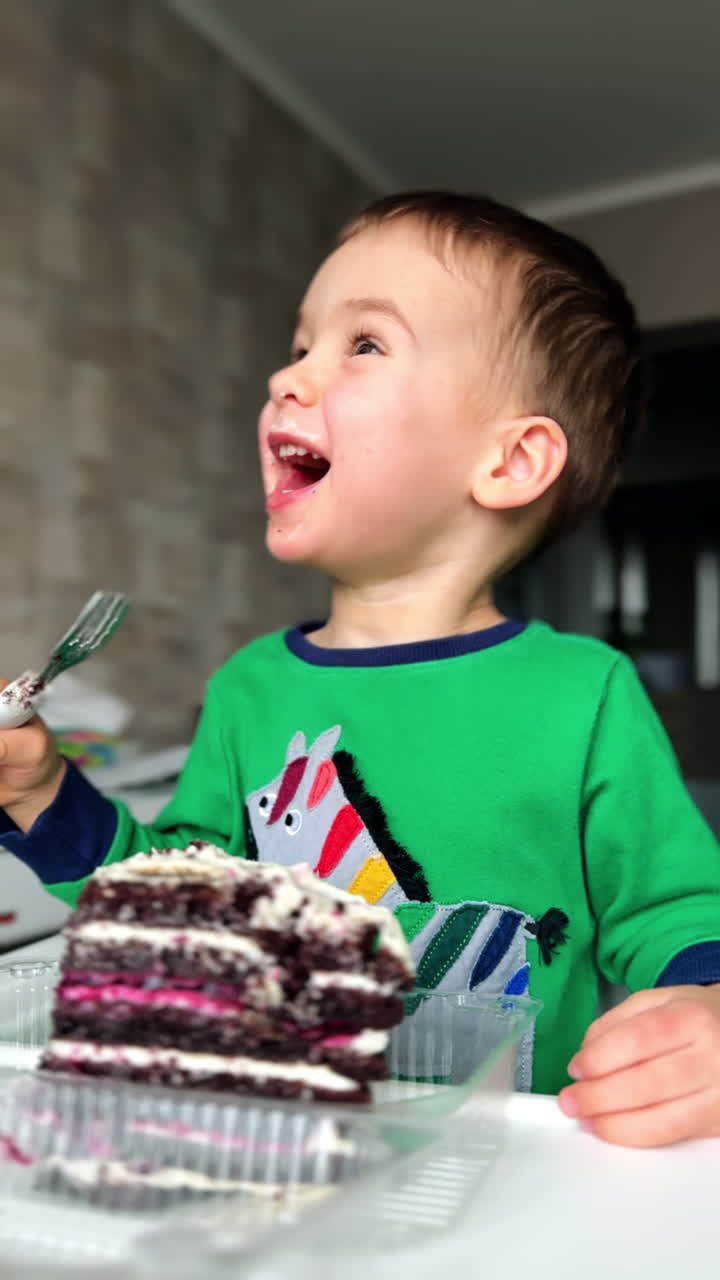 Happy Caucasian toddler in green shirt eating the cake. Kid deeps his fork into the big piece of dessert, licks the fork and laughs cheerfully. Vertical video.