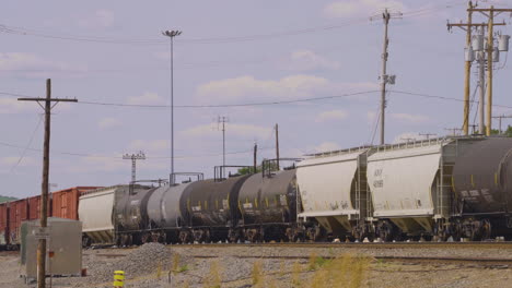 a train comprised of box cars, tanker cars and hopper cars moves across the railroad tracks surrounded by utility poles