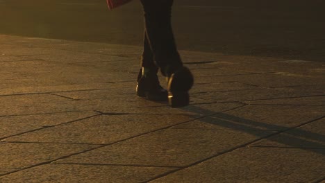 woman in stylish boots confidently walking along stone pavement, casting long shadows during golden sunset light, capturing urban evening atmosphere