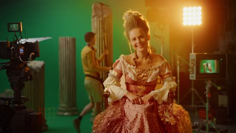 actriz sonriente con un vestido renacentista, sentada en una silla mirando a la cámara. pantalla verde en el fondo. estudio de rodaje historia blockbuster. en el estudio de cine período de traje drama set de película