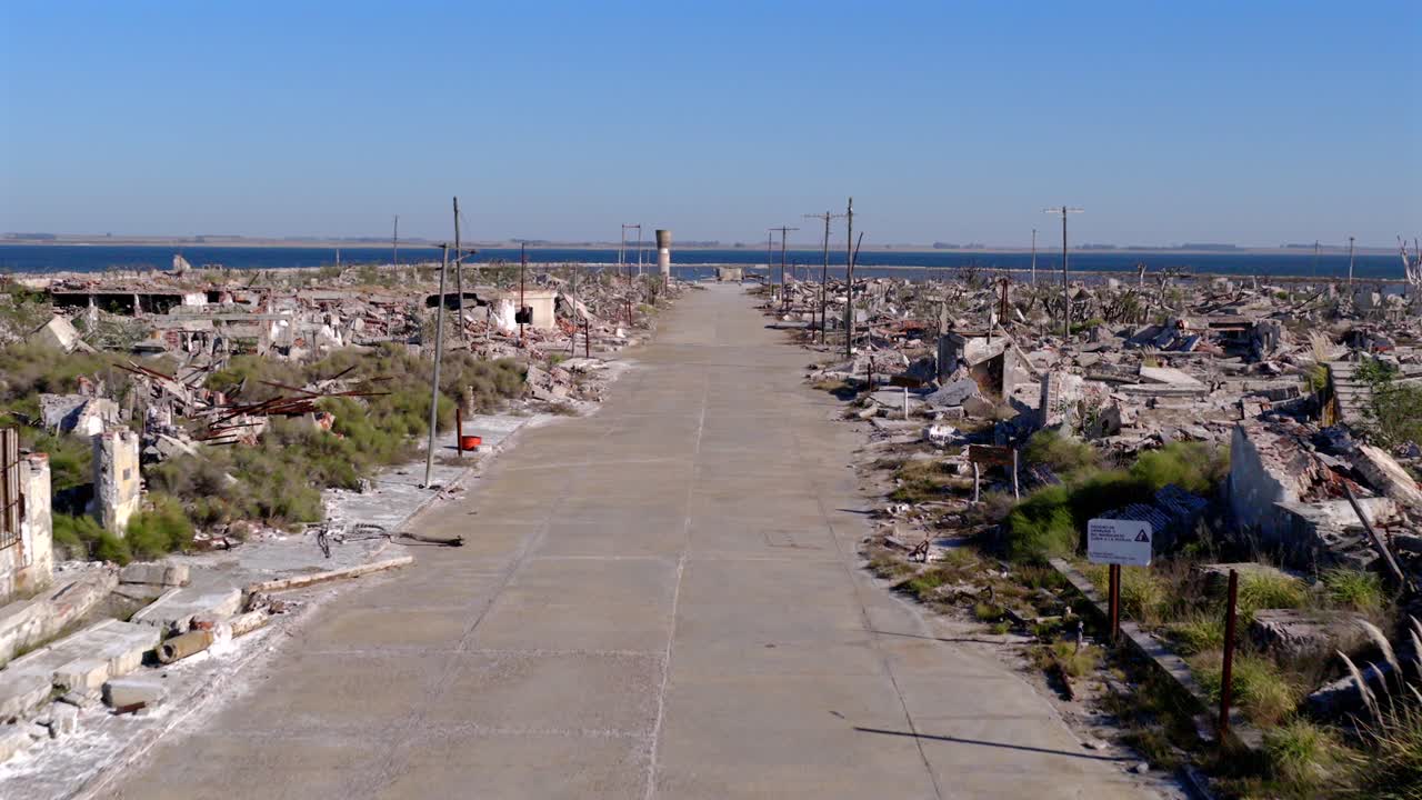 Drone establishing shot tracks Villa Epecuén’s broad central roadway, bordered by the battered remains of buildings destroyed by the flood of 1985, with crumbling walls and scattered vegetation