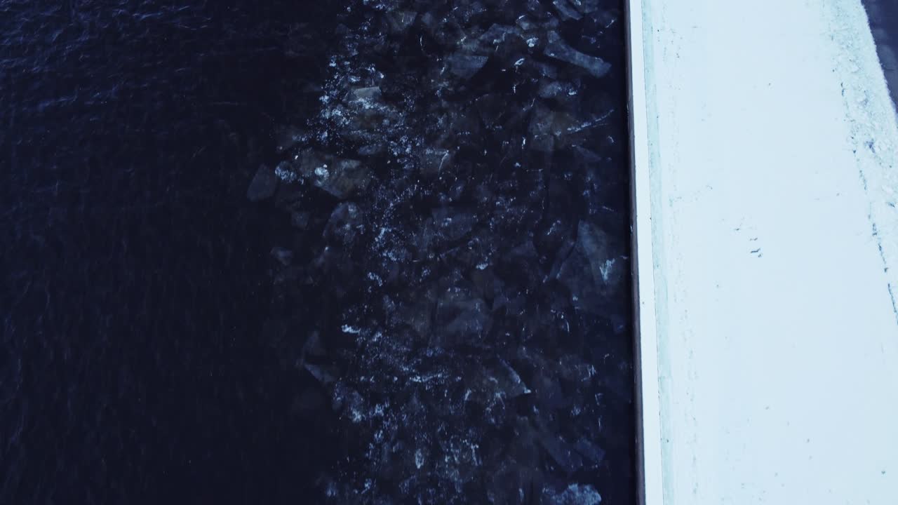 Drifting ice blocks rinse in shore of a dark deep river in winter, aerial top down view