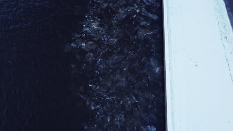 drifting ice blocks rinse in shore of a dark deep river in winter, aerial top down view