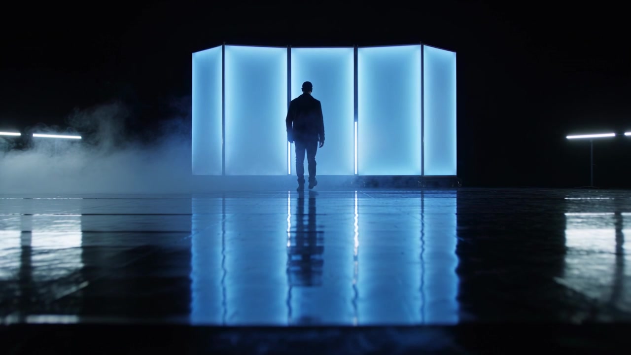 Uomo che cammina con giacca scura, si muove verso pannelli luminosi in uno studio, attratto da una luce blu.