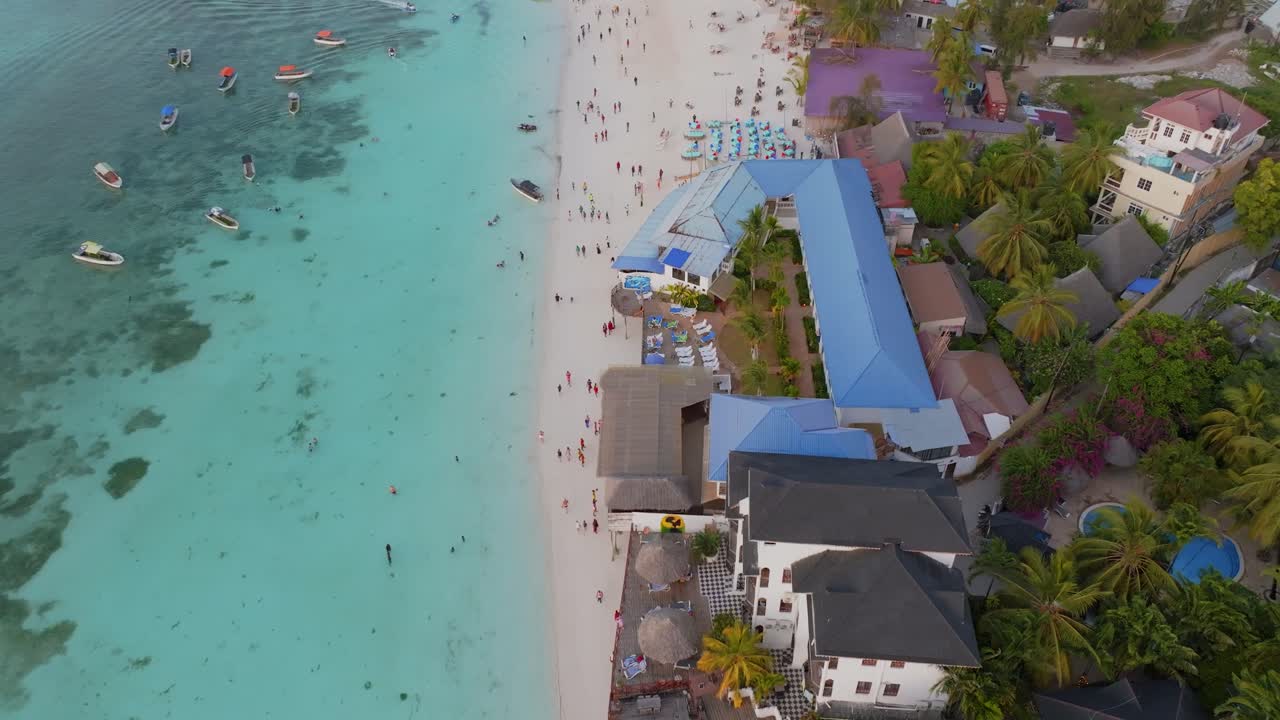 Ripresa cinematografica dall'alto con drone sopra la spiaggia di Nungwi a Zanzibar, che mostra persone che si rilassano e ristoranti sul mare lungo la costa. L'acqua turchese, la sabbia bianca e l'energia tropicale creano un'atmosfera vivace