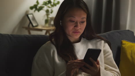 woman spending evening at home on sofa wearing wireless earbuds and streaming music or podcast from mobile phone 1