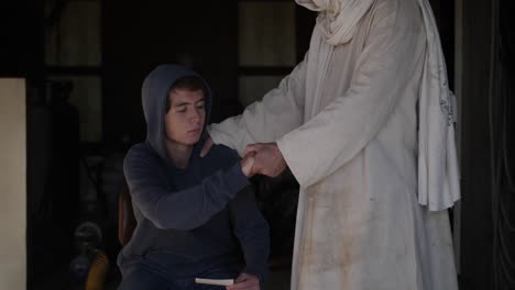 Jesus,-wearing-a-white,-tattered-robe,-takes-the-hand-of-a-young,-sad,-teenage-boy-and-helps-him-to-stand-up