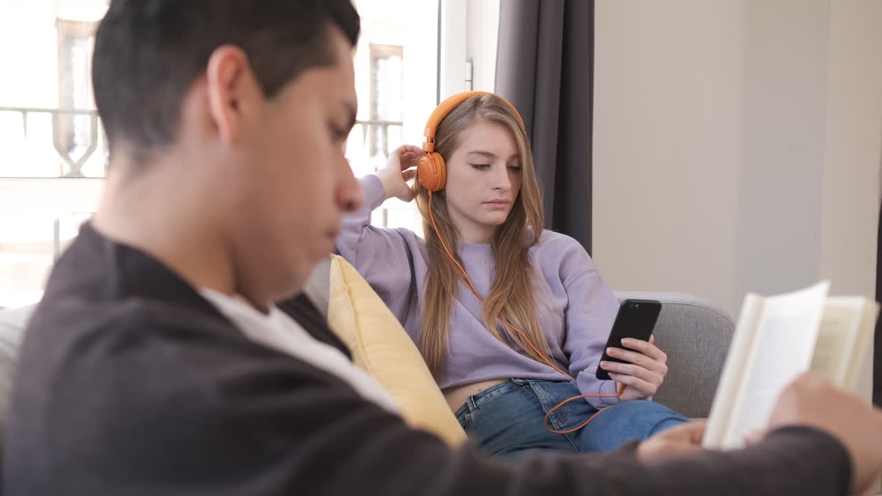 Young couple relaxing and sitting on couch at home while reading a book and listening music with headphones