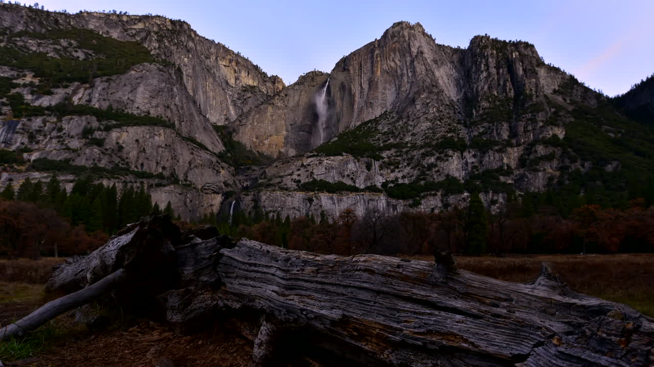 Sunrise time lapse with a log in the foreground and a picturesque Yosemite Falls waterfall in the distance on a granite cliff