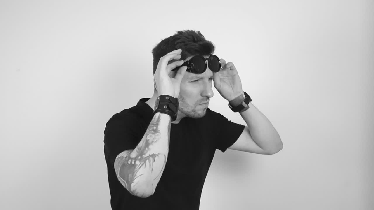 Grayscale footage of a tattooed man with short hair and short beard wearing black rounded steampunk retro vintage sunglasses, lifting them up and leaning forward to look at something a bit closer.