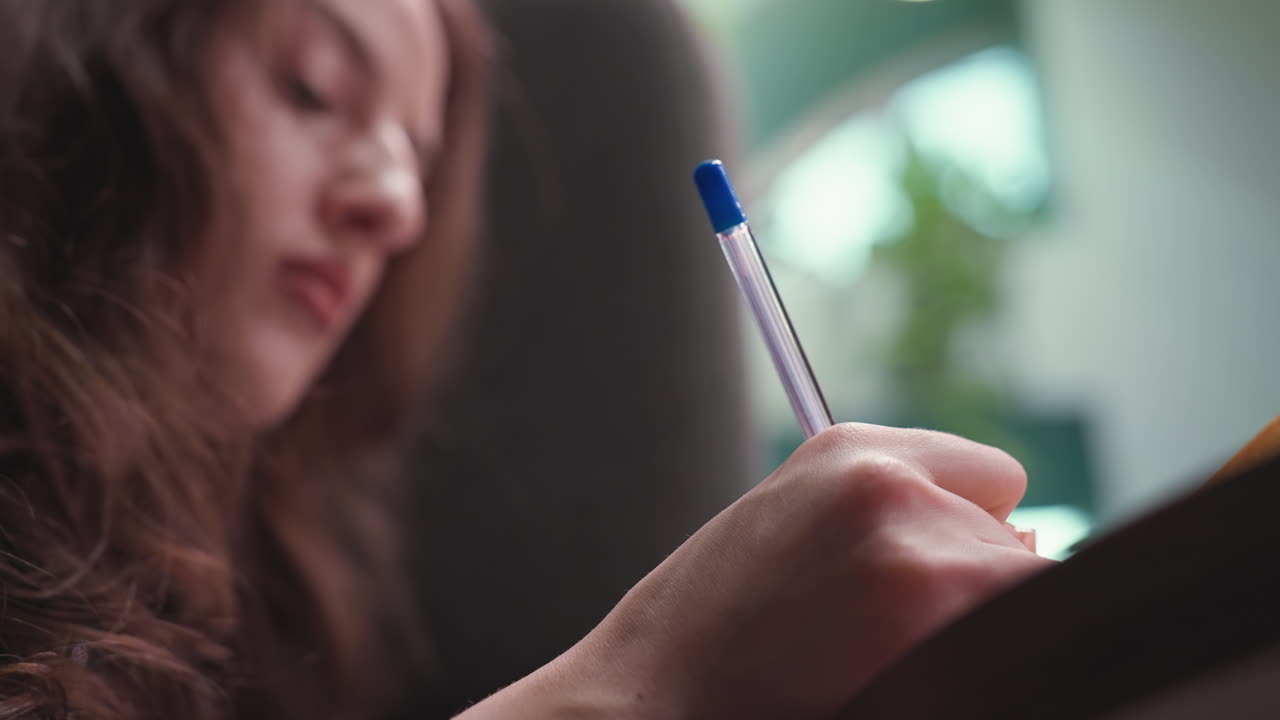 Close up view of woman concentrating while writing with blue pen in notebook, soft lighting and blurred indoor background add calm, thoughtful mood perfect for themes of study, planning, or reflection