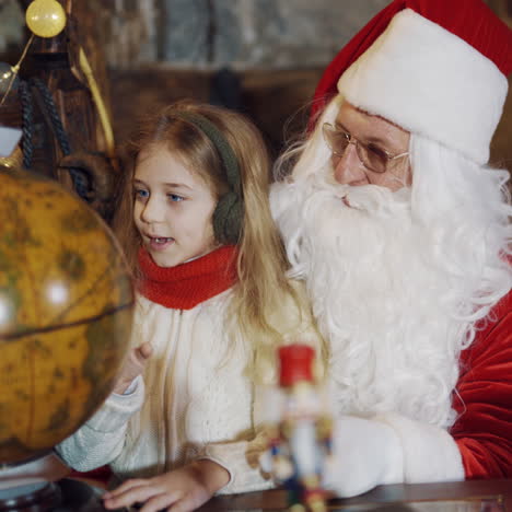 pretty little girl sitting with santa claus in front of the globe model. santa in red and white suit looking at globe together with a child at christmas.