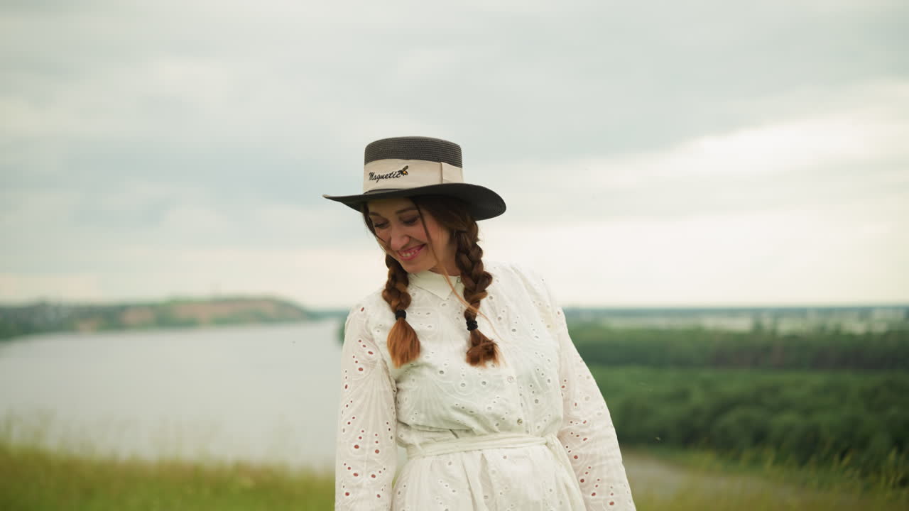 una mujer está de pie en un campo de hierba verde con un lago visible en el fondo. está usando un sombrero elegante y un vestido blanco con patrones intrincados, haciendo una cara graciosa mientras mira hacia el lado