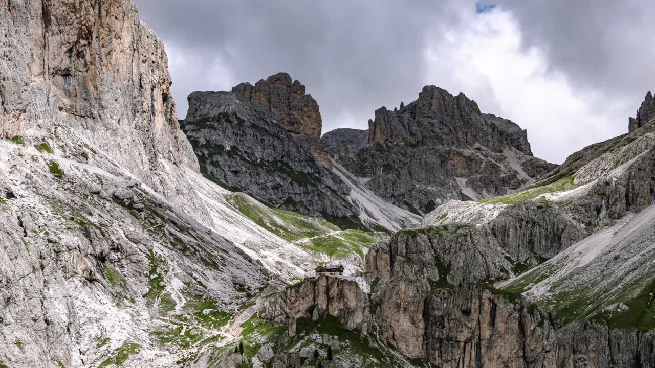 Vista aerea in hyperlapse della zona montuosa del Santner nelle Dolomiti, che mostra aspre scogliere calcaree, un rifugio alpino remoto e cieli nuvolosi drammatici