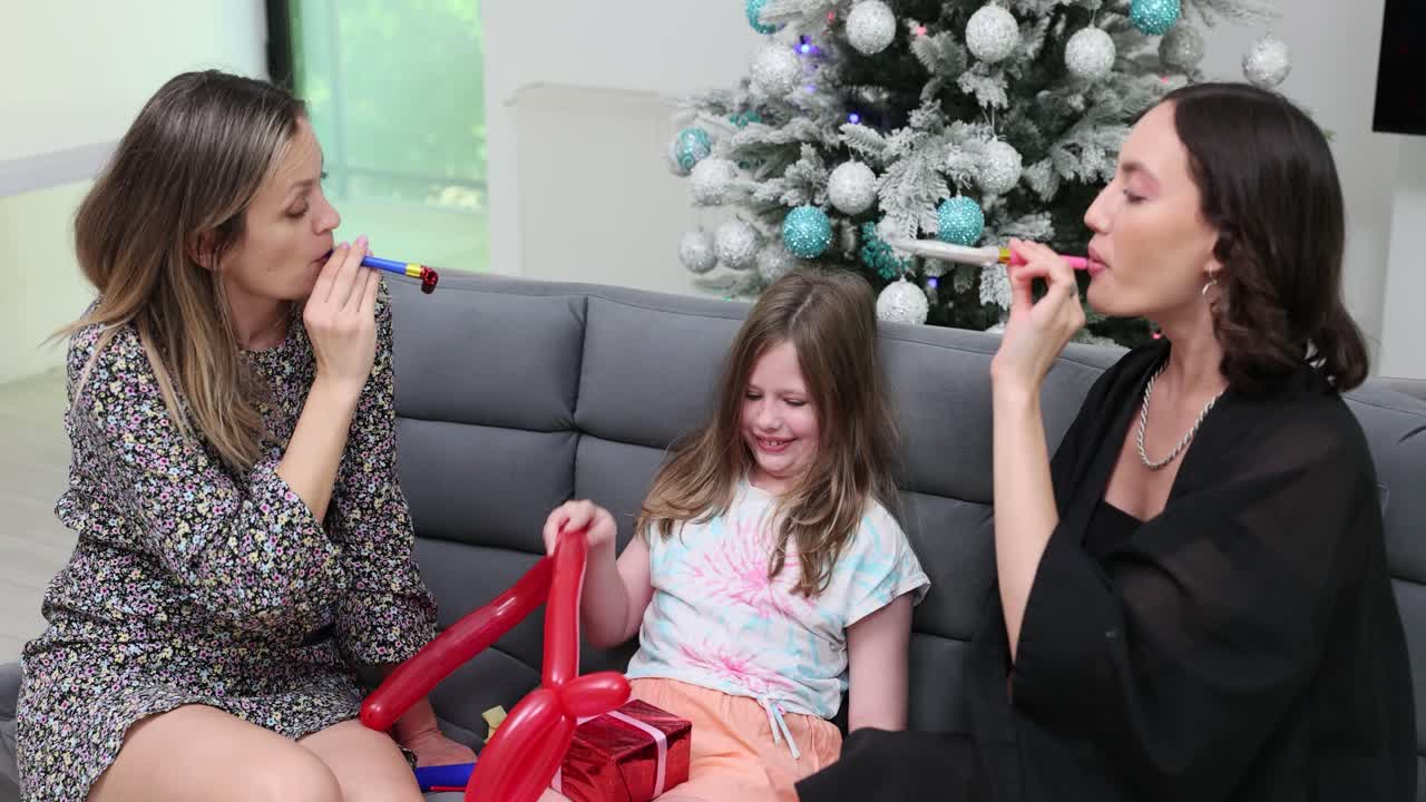 Three women and a girl celebrate a holiday with gifts and noisemakers by a Christmas tree