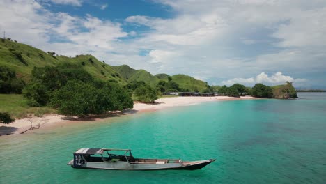 aerial shot over the stunning emerald sea and pink-hued beach on the exotic island of the komodo archipelago, with green hills as a backdrop under a bright blue sky with soft white clouds.