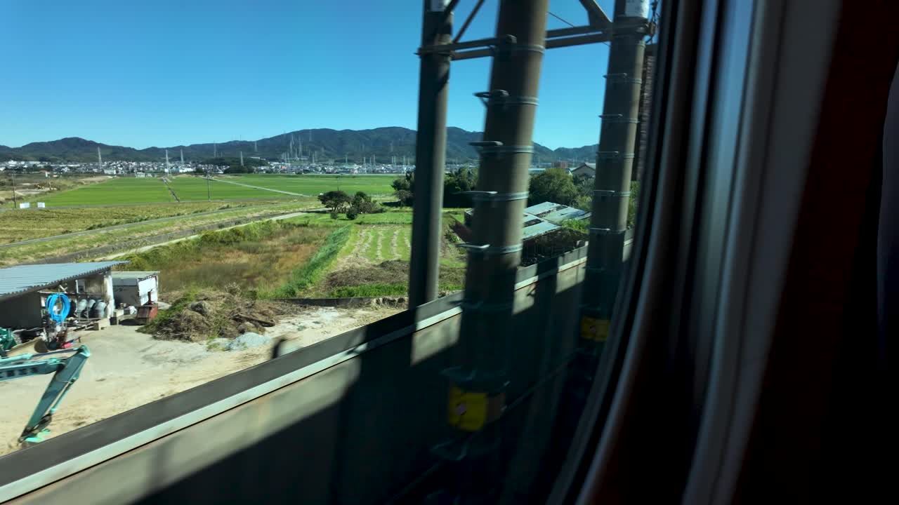 la vista panorámica de los campos, árboles y montañas del campo japonés, tomada a través de la ventana de un tren en movimiento, el cielo despejado y el paisaje vibrante evocan una experiencia de viaje tranquila y pintoresca.