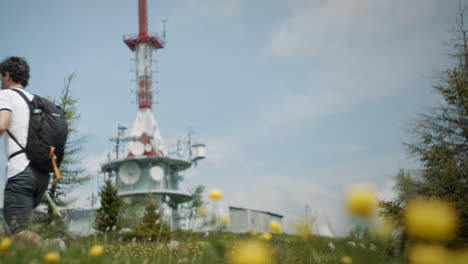 Excursionista-Caminando-Con-Bastones-De-Senderismo-Más-Allá-De-La-Cámara-En-Primer-Plano-Hay-Flores-Amarillas-Y-En-El-Fondo-Está-La-Torre-De-Radio