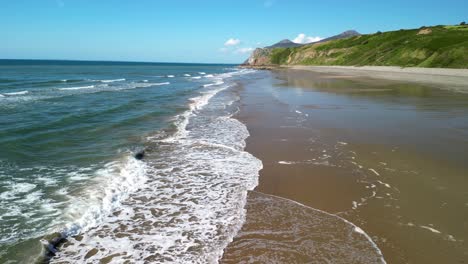 Nefyn-beach-fast-drone-low-level-sweep-over-waves-breaking,-beautiful-North-Wales-coastline,-UK