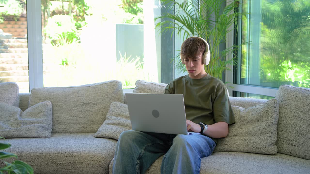 Youth male looking down at laptop, typing and adjusting posture while working at home, copy space