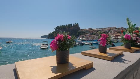 Trucking-shot-of-boats-floating-on-idyllic-Ionian-coastline-from-behind-flower-arrangements,-Parga,-Greece