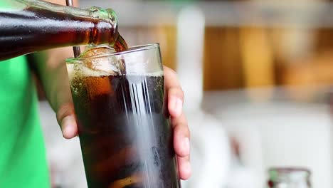 A person in a green shirt pours soda from a bottle into a glass filled with ice.
