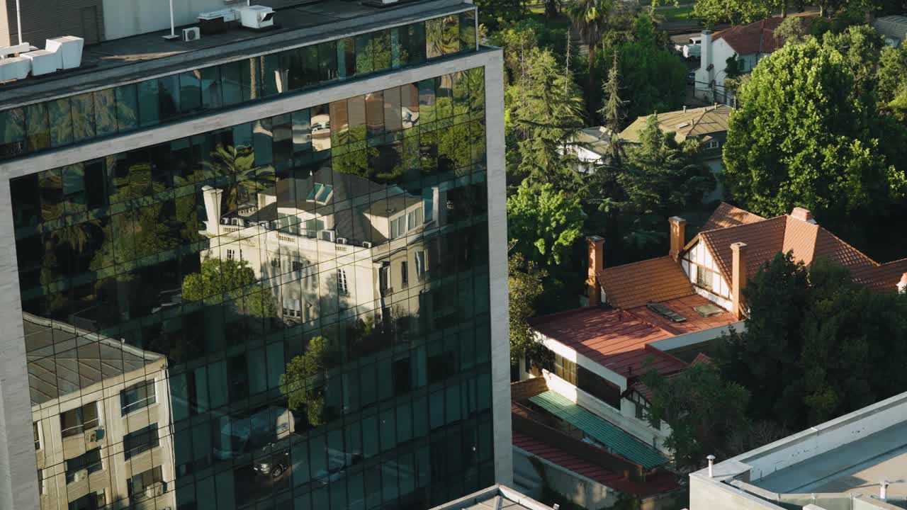 Prédio de escritórios moderno de vidro em Providencia, Santiago do Chile, refletindo as árvores e casas próximas, com ruas residenciais arborizadas e telhados ao fundo