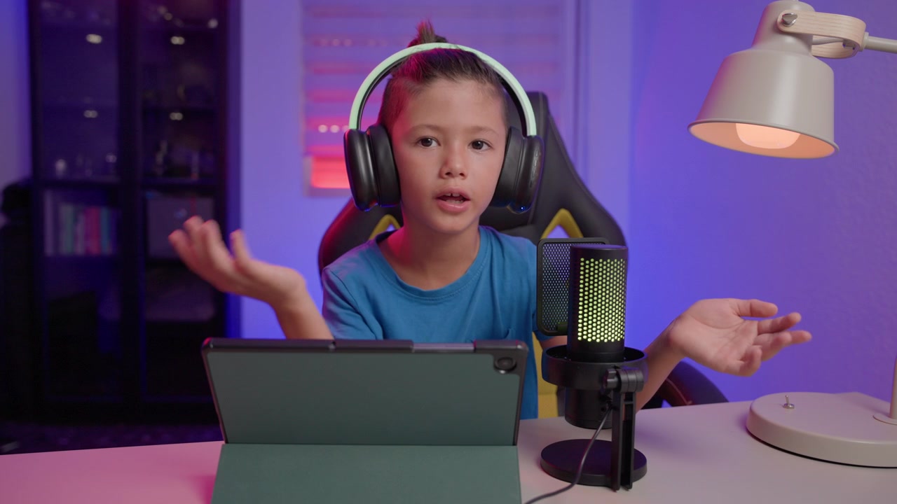 Un giovane influencer che indossa cuffie parla e gesticola alla telecamera davanti a un microfono e un tablet, illuminato da luci ambientali colorate.