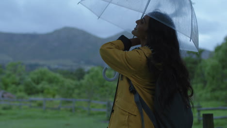 attractive young indian woman holding umbrella smiling happy prepared enjoying calm rainy day in vibrant green countryside farm background