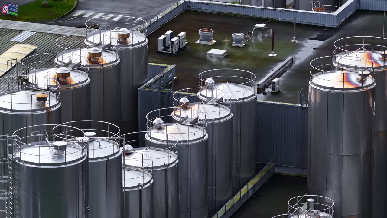 Aerial close-up of stainless steel storage tanks and silos on an industrial rooftop, with steam venting and equipment visible. Modern manufacturing infrastructure and energy concept