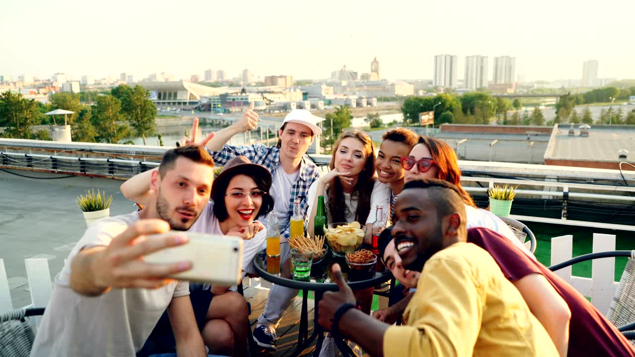 행복한 젊은 남녀들은 스마트폰으로 셀피를 찍으며 포즈를 취하고, 웃고, 손짓을 하며, 식탁 위에서 음식과 음료와 함께 휴일을 축하하고 있습니다.