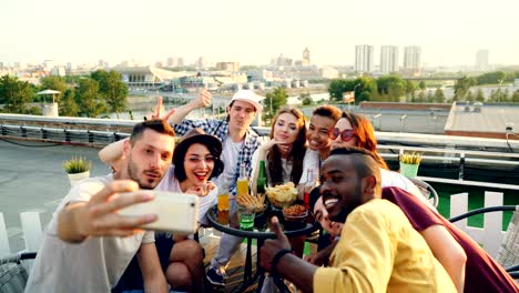 hombres y mujeres jóvenes felices se están tomando selfies con teléfonos inteligentes posando, riendo y haciendo gestos con las manos celebrando la fiesta en el techo en la mesa con comida y bebidas.