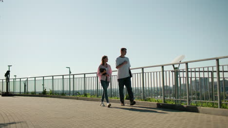 lady in pink shirt holding notebook walking alongside brother on paved path near iron railing, engaging in cheerful conversation surrounded by greenery, cityscape, and urban architecture