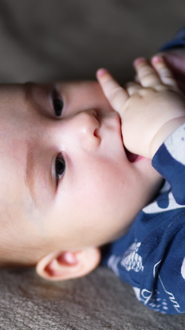 Calm peaceful baby lies on bed on his back. Healthy Caucasian child looks up and holds his fingers in mouth. Grey backdrop. Vertical video