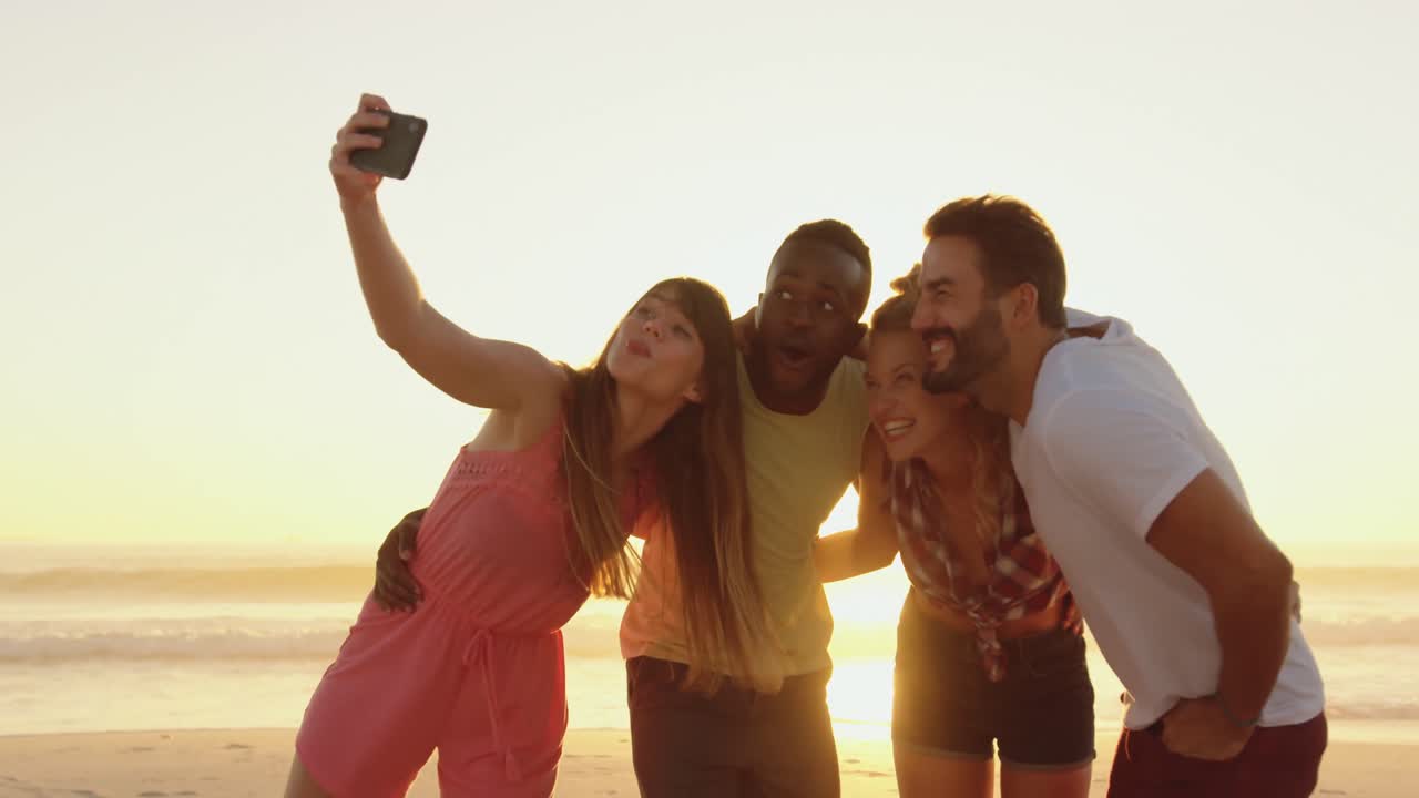 Amici giovani che si fanno selfie in spiaggia al tramonto 4k
