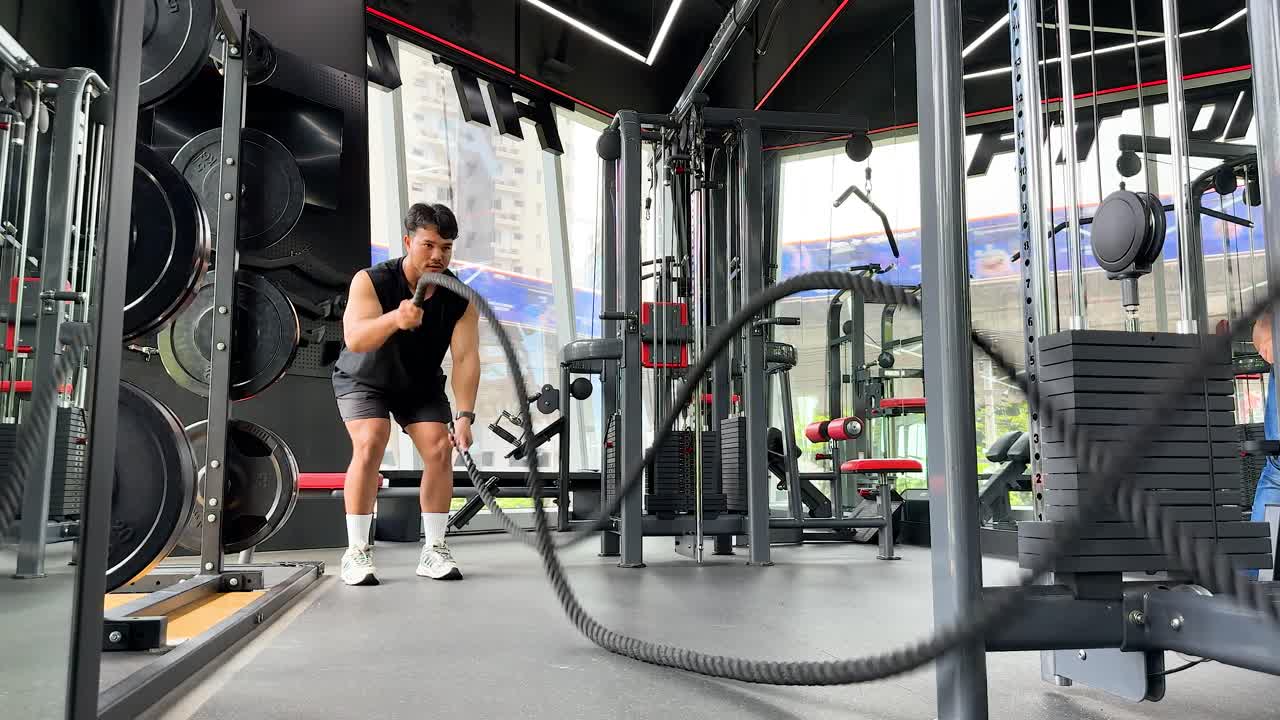 A person performs a dynamic battle rope exercise in a well-equipped gym with bright lighting and modern equipment