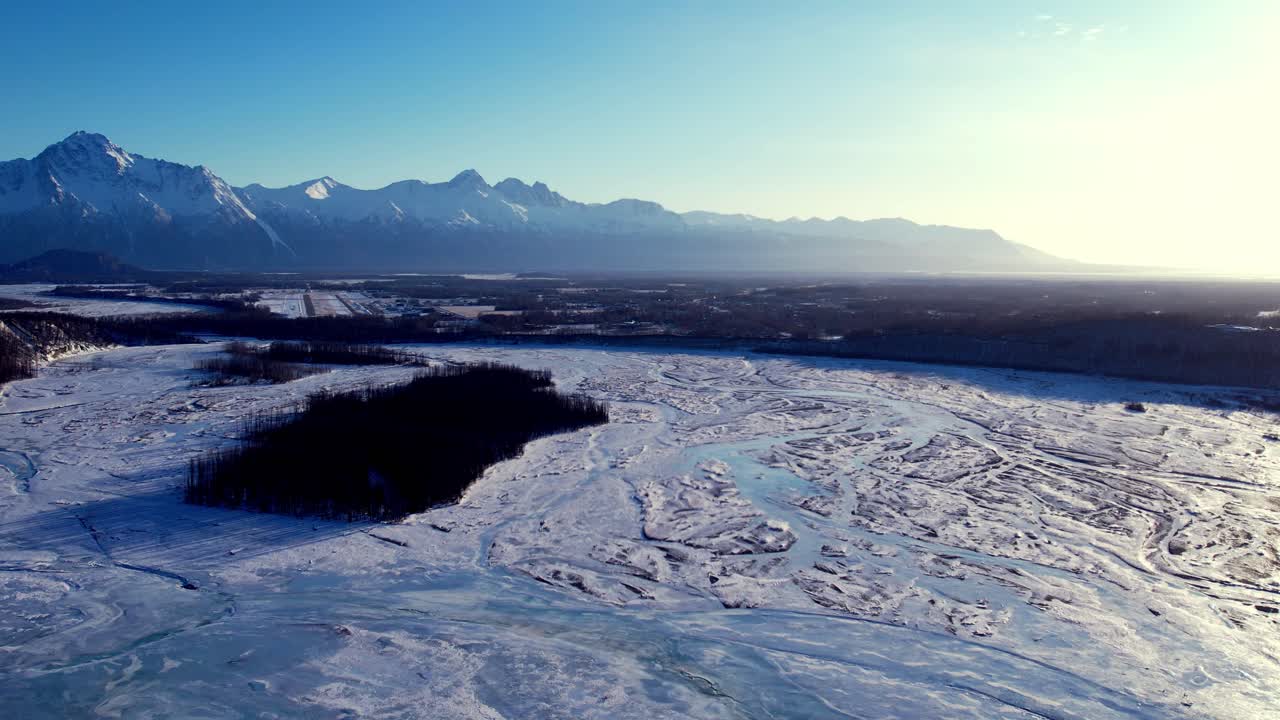 Droneflyvning i mellemlav højde mod vintersolen over den store frosne Matanuska-flod med flettede flodarme nær Palmer, AK. En isoleret træklædt ø bryder isfladen, tågede bjergkæder gløder i bagbelyst gyldent lys