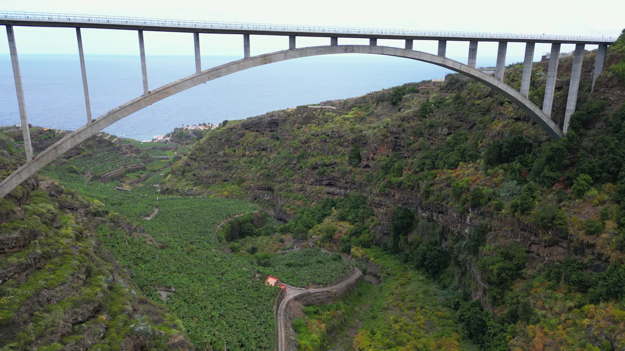 A large concrete bridge spans a lush green valley filled with banana plantations, with the ocean visible in the background.