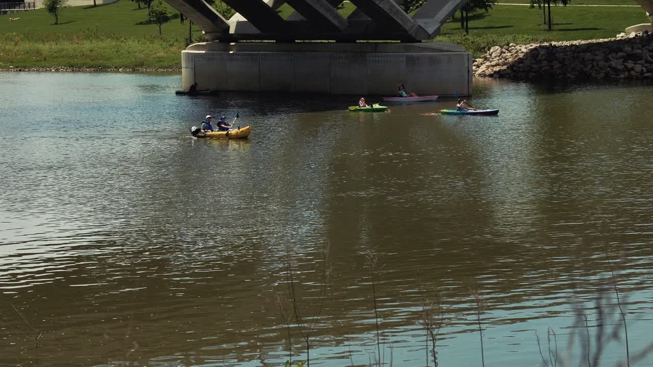 Rising above green grass by river's edge to reveal kayakers paddling downstream under bridge