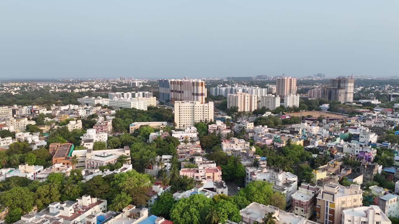 Video aereo di Jones Rd, Periyapet, Saidapet, Chennai. Un grattacielo colorato spicca tra un mix di edifici nuovi e vecchi. Un binario ferroviario si estende sotto, mentre si intravedono un campo lontano e lavori di costruzione in corso.