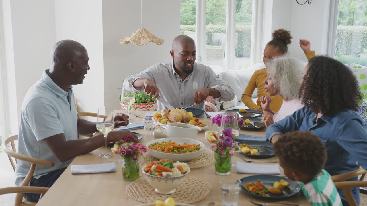 Père découpant (la viande) pendant que la famille multigénérationnelle est assise autour de la table à la maison et apprécie le repas