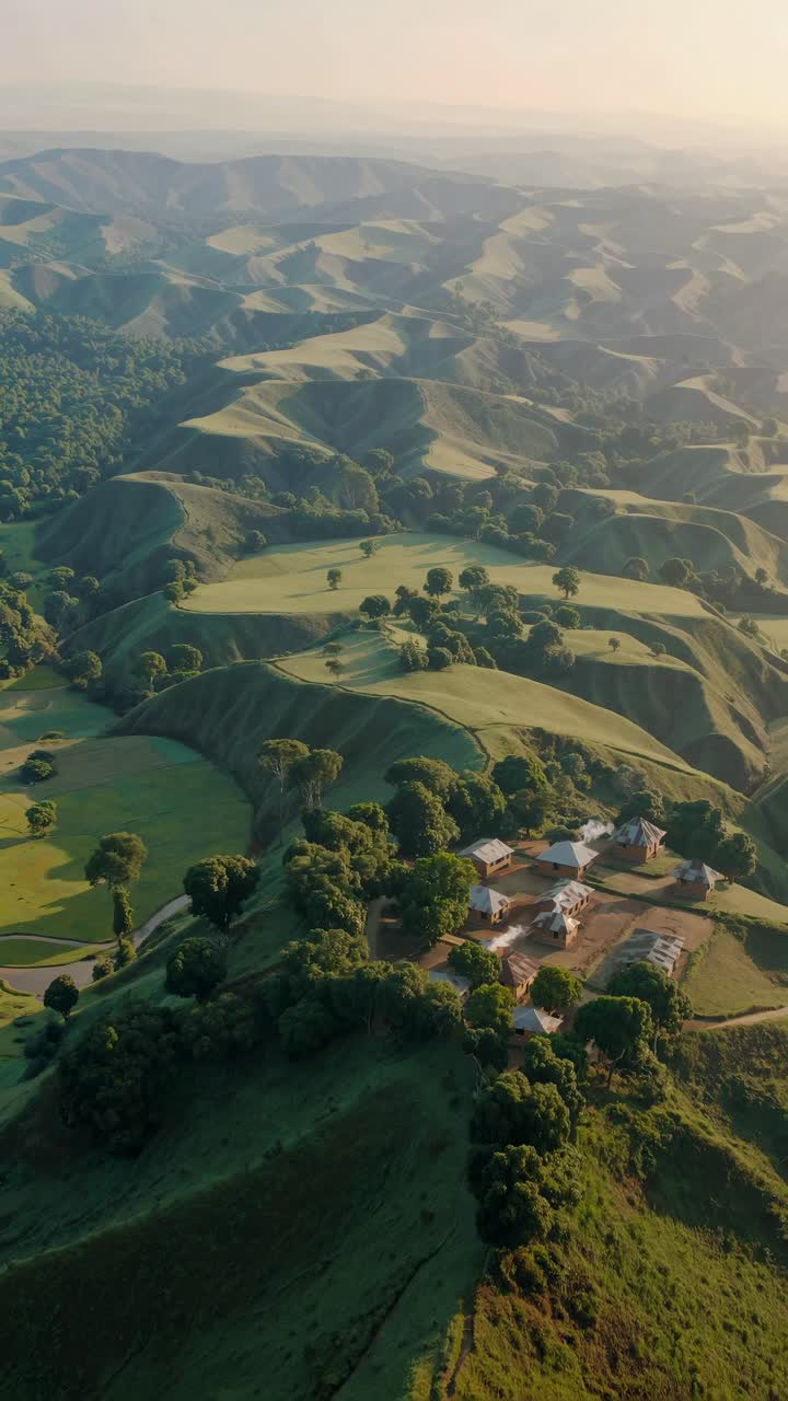 Aerial view of lush green hills with scattered trees and a small village, captured in a cinematic