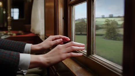 hands resting on a classic wooden window sill, looking out at a scenic green countryside view