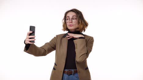 Hermosa-Mujer-Joven-Con-Chaqueta-Marrón-Preparándose,-Mirando-Su-Teléfono-Inteligente-Y-Tomándose-Una-Selfie-Aislada-En-Blanco