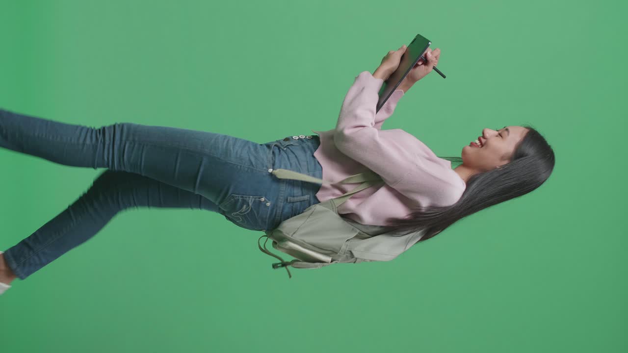 vista lateral de una estudiante asiática escribiendo en una tableta y caminando en un estudio de pantalla verde