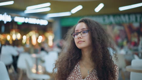 dama con cabello rizado vintage y gafas sentada en el restaurante, mirando pensativo a su alrededor, el fondo muestra luces bokeh borrosas y otras personas, creando una escena acogedora y ambiental
