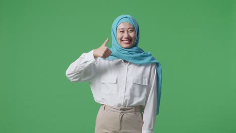 아시아 여성이 미소 짓고 엄지손가락을 올리며 녹색 스크린 배경 스튜디오에 서서 카메라를 향해 몸짓을 합니다.