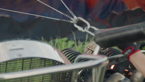 close up of person preparing hot air balloon burner setup as pilot ignites engine flame through metal hoses and gauges during preflight inflation sequence on grassy field under cloudy sky
